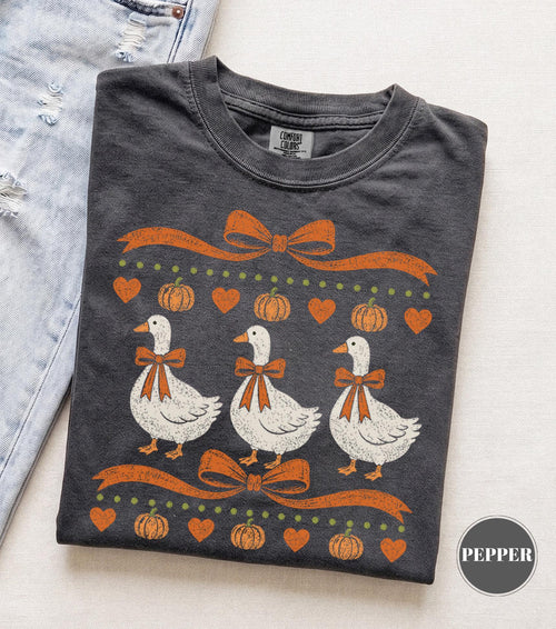 May include: A charcoal gray t-shirt featuring a graphic print of three white geese wearing orange bows. The design includes orange ribbons, small orange hearts, and orange pumpkins arranged in rows. The shirt has a crew neckline and a Comfort Colors label.