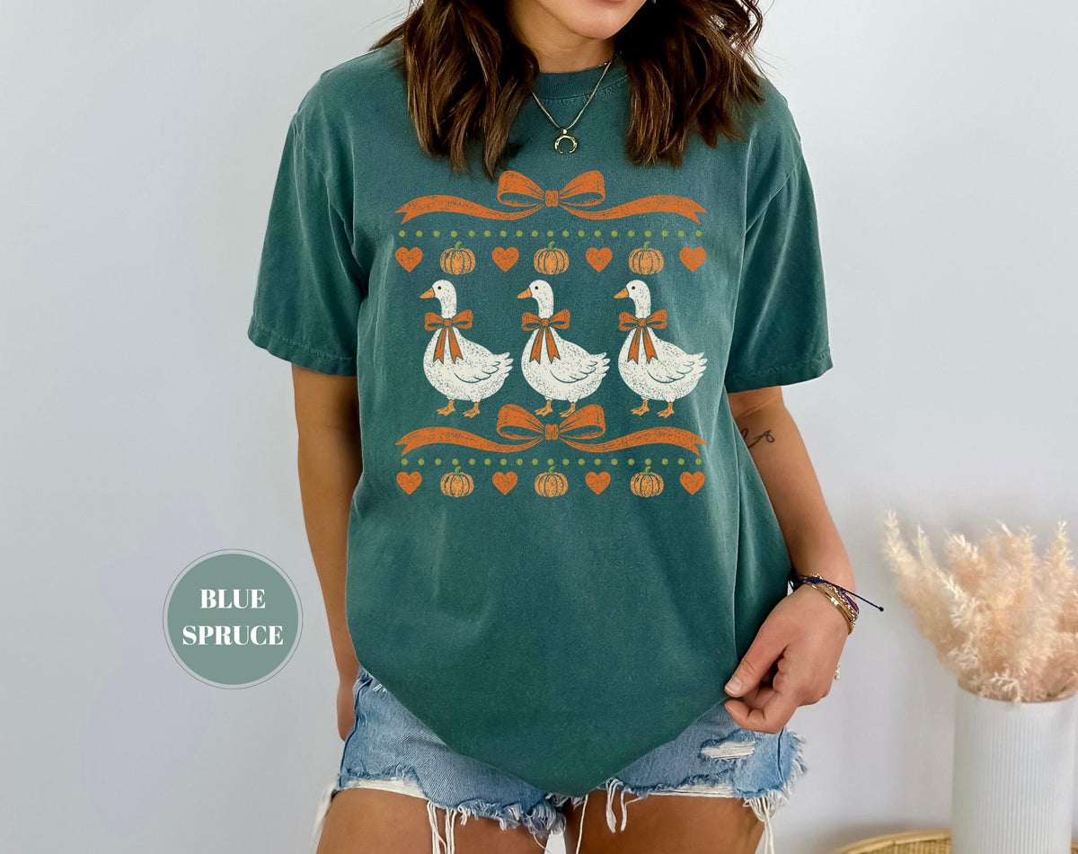 May include: A blue spruce colored short-sleeve t-shirt featuring a graphic print of three white geese wearing orange bows. The design includes orange ribbons, small orange pumpkins, and orange hearts arranged in horizontal rows across the chest.