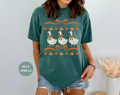 May include: A blue spruce colored short-sleeve t-shirt featuring a graphic print of three white geese wearing orange bows. The design includes orange ribbons, small orange pumpkins, and orange hearts arranged in horizontal rows across the chest.