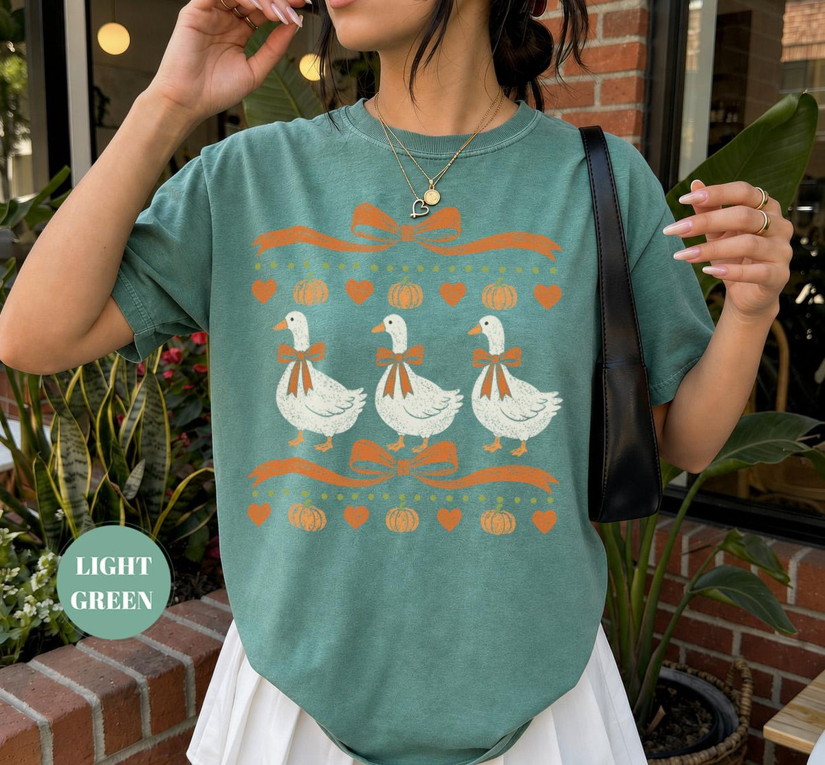 May include: A light green short-sleeve t-shirt featuring a graphic print of three white geese wearing orange bows. The design includes orange ribbons, small orange pumpkins, and orange hearts arranged in horizontal rows across the chest.