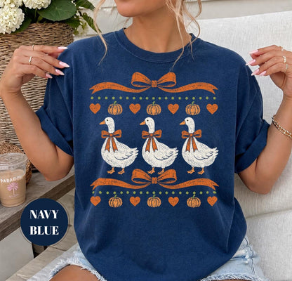 May include: A navy blue t-shirt featuring a graphic print of three white geese wearing orange bows. The design includes orange ribbons, small orange pumpkins, and orange hearts arranged in horizontal rows across the front of the garment.