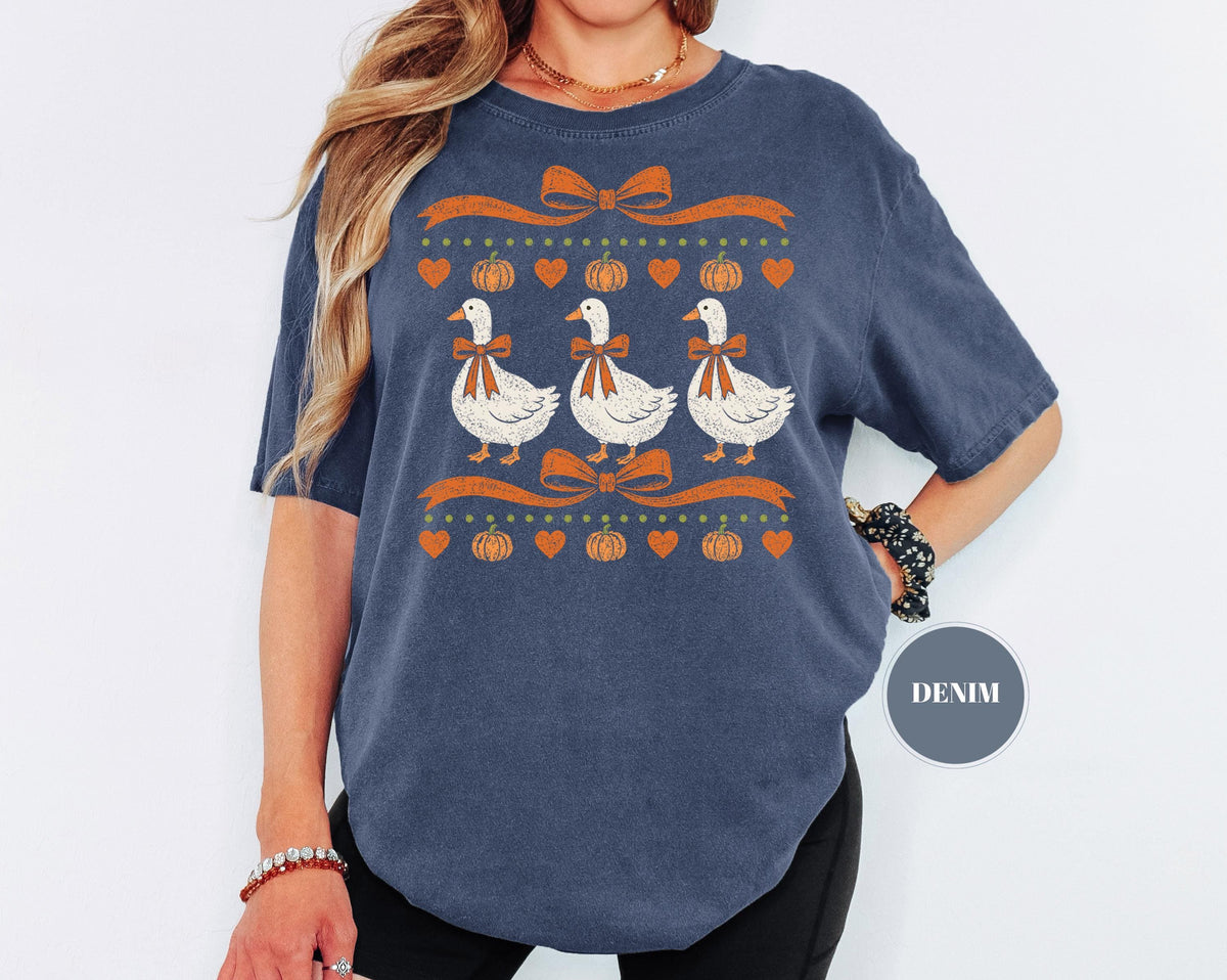 May include: A denim blue short-sleeve t-shirt featuring a graphic print of three white geese wearing orange bows. The design includes orange ribbons, small orange pumpkins, and orange hearts arranged in rows above and below the geese.