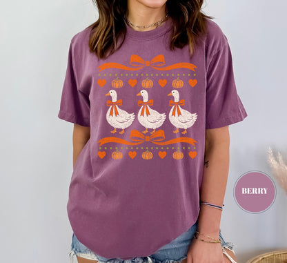 May include: A berry-colored short-sleeve t-shirt featuring a graphic print of three white geese wearing orange bows. The design includes orange ribbons, pumpkins, and heart shapes arranged in rows. The word BERRY appears in a small circle.