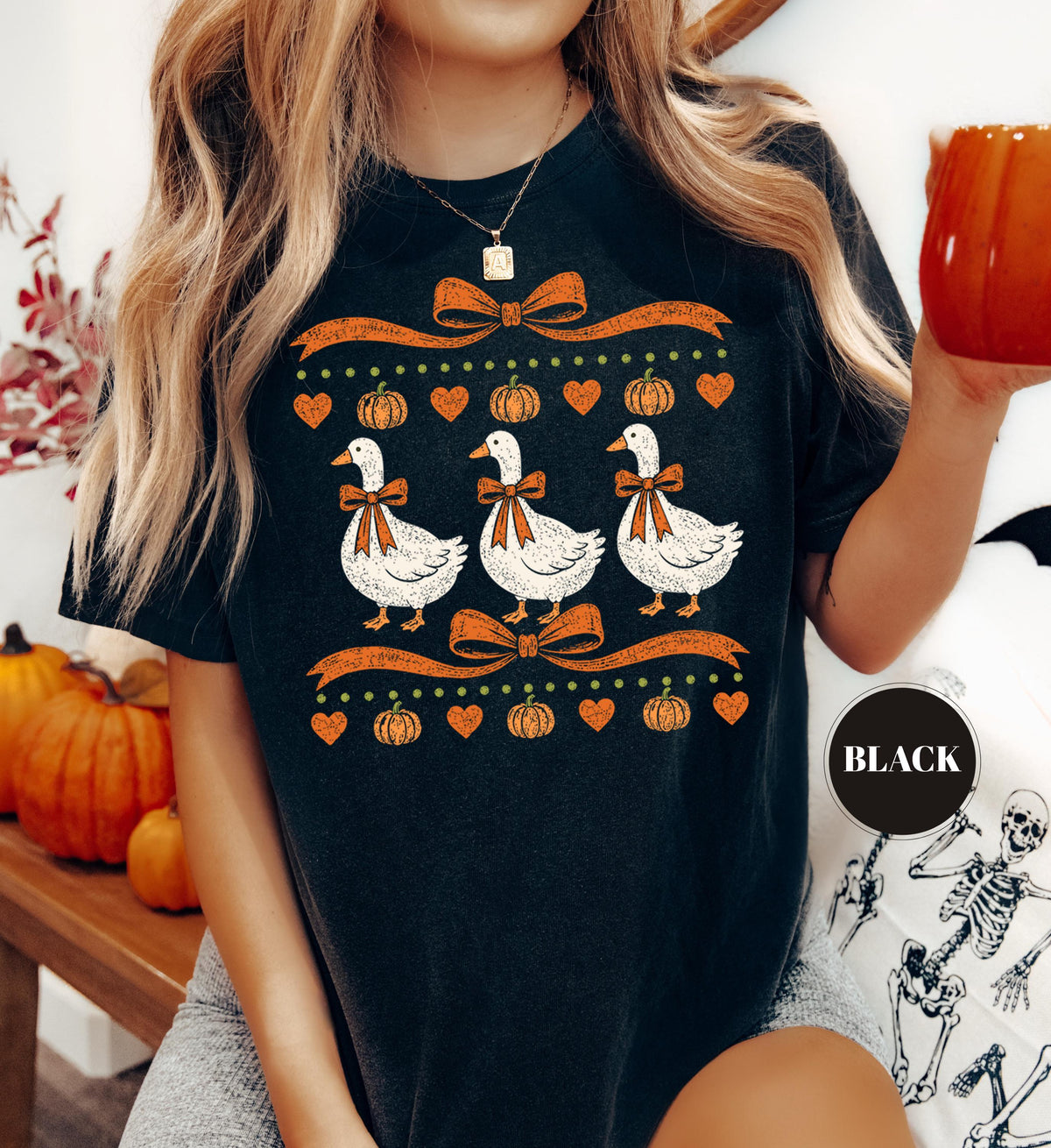 May include: A black t-shirt featuring a graphic print of three white geese wearing orange bows, surrounded by orange pumpkins, orange hearts, and orange ribbons. The design is arranged in a horizontal pattern with a small black circle label.