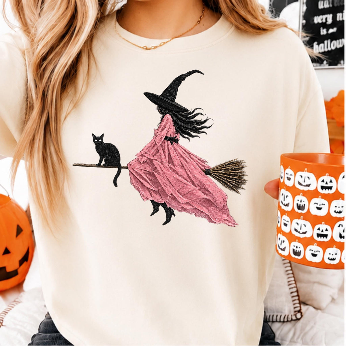 May include: A cream-colored crewneck sweatshirt featuring a graphic print of a figure in a long pink dress and a black pointed hat riding a broomstick, accompanied by a black cat. The design is centered on the front of the garment.