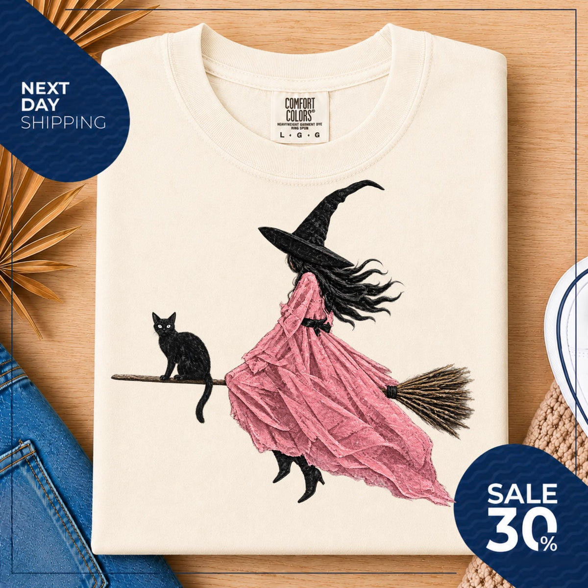 May include: A cream-colored t-shirt featuring a graphic illustration of a figure in a pointed hat and a flowing pink dress riding a broomstick, accompanied by a black cat. The shirt has a Comfort Colors brand label at the neck with size L.