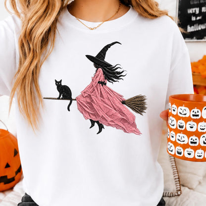 May include: A white crewneck sweatshirt featuring a graphic illustration of a witch wearing a black hat and a flowing pink dress, riding a broomstick with a black cat perched on the handle. The design is centered on the front of the garment.