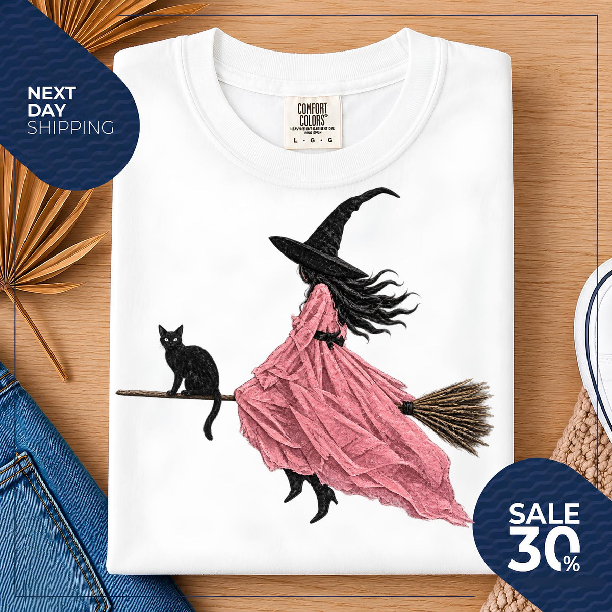 May include: A white t-shirt featuring a graphic illustration of a figure wearing a black witch hat and a flowing pink dress, riding a broomstick with a black cat sitting on the handle. The shirt has a Comfort Colors brand label at the neck.