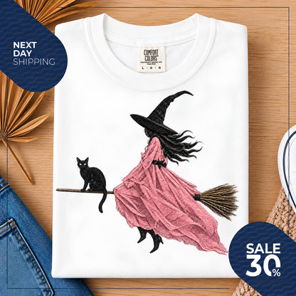 May include: A white t-shirt featuring a graphic illustration of a figure wearing a black witch hat and a flowing pink dress, riding a broomstick with a black cat sitting on the handle. The shirt has a Comfort Colors brand label at the neck.