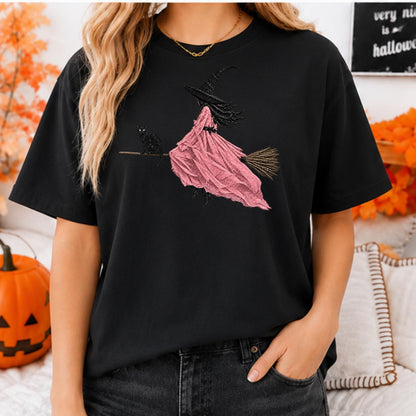 May include: A black short-sleeve t-shirt featuring a graphic print of a witch wearing a pink dress and a black hat, riding a broomstick with a black cat. The design includes a textured pink gown and a brown broom with visible bristles.