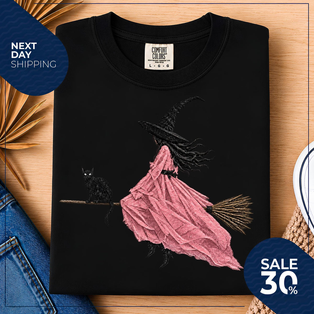 May include: A black crewneck t-shirt featuring a graphic print of a witch in a long pink dress and a pointed hat riding a broomstick, accompanied by a black cat. The shirt has a Comfort Colors brand label at the inner neckline showing size L.