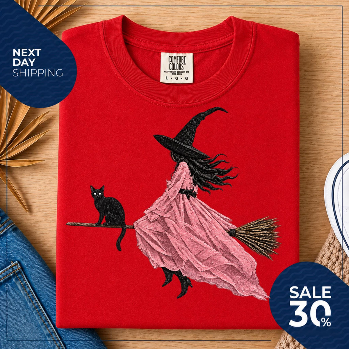 May include: A red crew neck t-shirt featuring a graphic illustration of a figure in a black witch hat and a flowing pink dress riding a broomstick, accompanied by a black cat. The inner neck label displays the text Comfort Colors L G G.