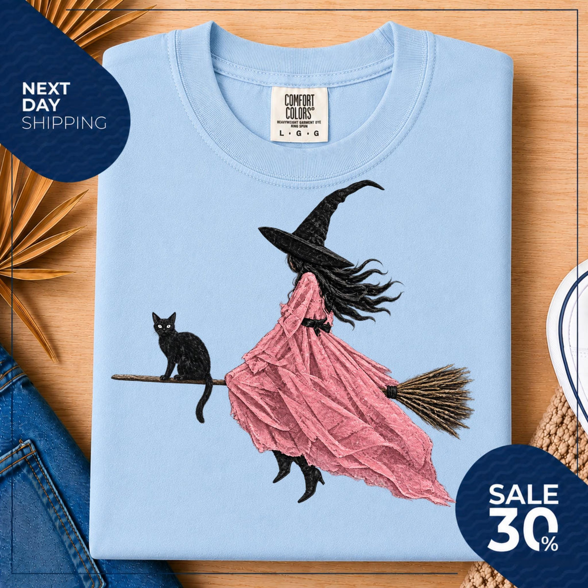 May include: A light blue crew neck t-shirt featuring a graphic illustration of a witch wearing a black hat and a flowing pink dress, riding a broomstick with a black cat sitting on the handle. The shirt has a Comfort Colors brand label.