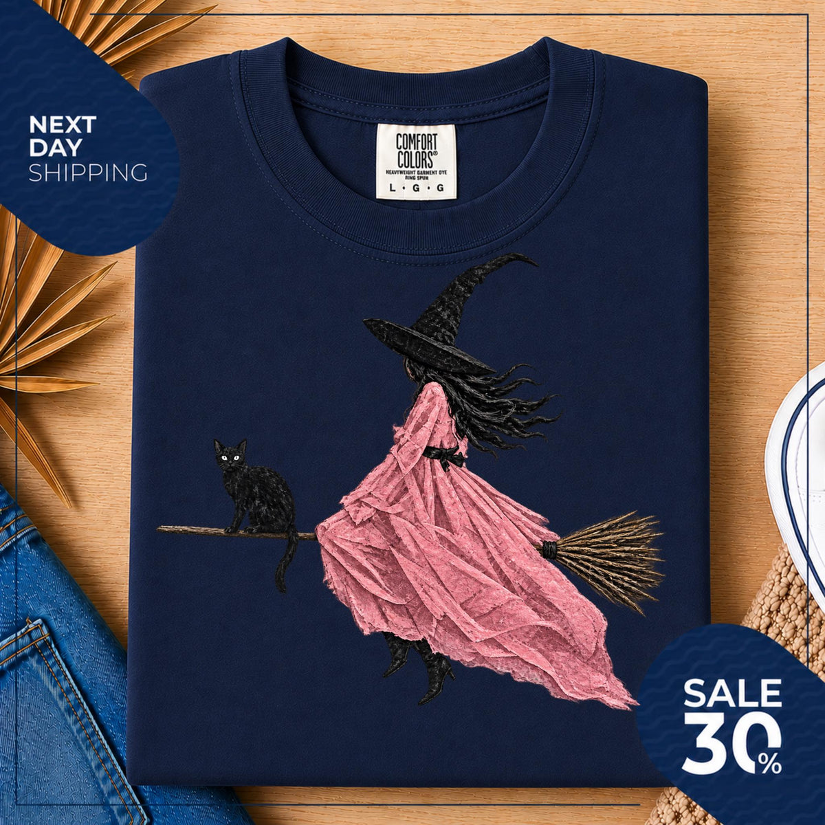 May include: A navy blue t-shirt featuring a graphic of a witch in a long pink dress and a black pointed hat riding a broomstick with a black cat sitting on the handle. The shirt has a Comfort Colors brand label at the neck showing size L.