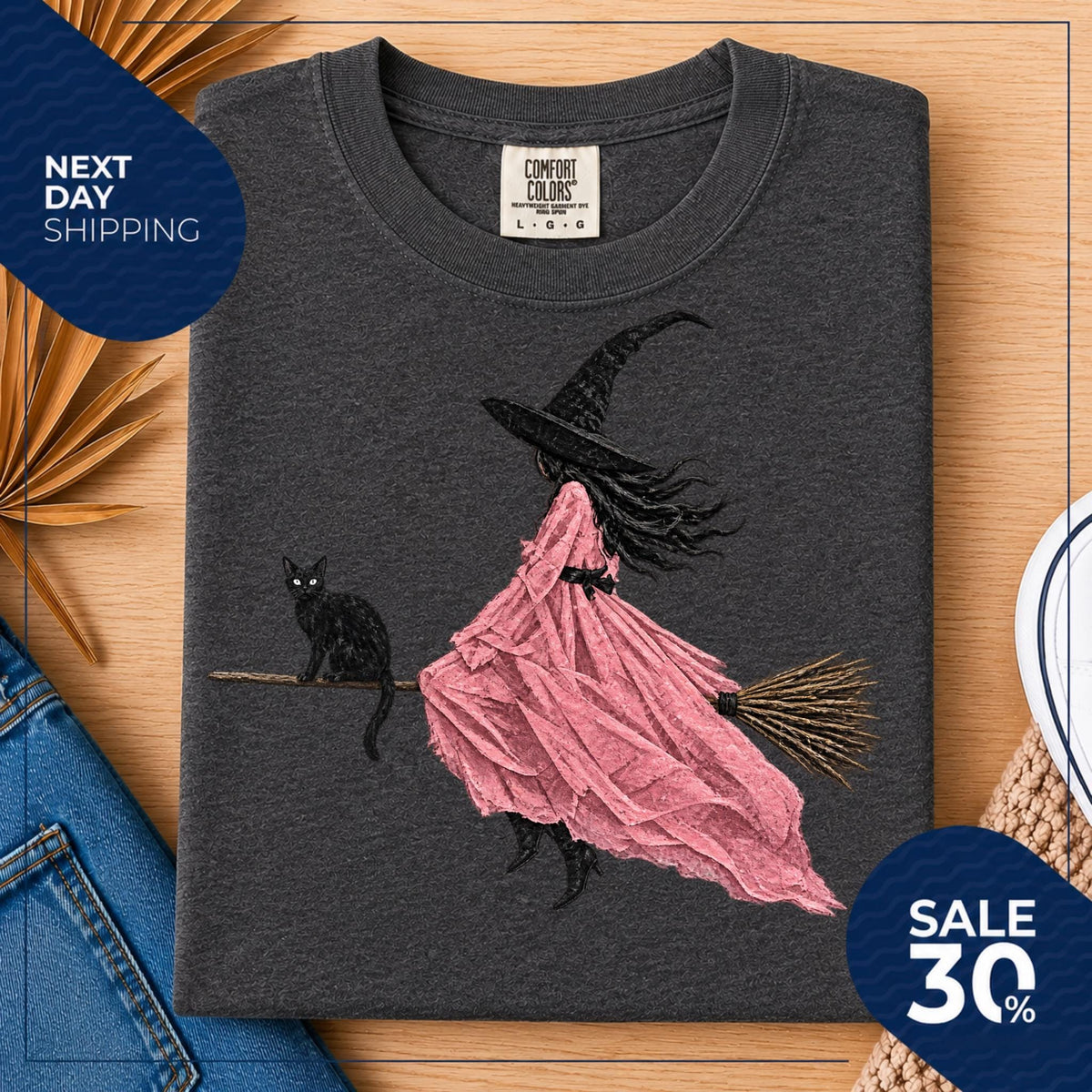 May include: A charcoal gray crew neck t-shirt featuring a graphic print of a figure in a black witch hat and a flowing pink dress riding a broomstick, accompanied by a black cat. The inner neck label reads Comfort Colors size L G G.