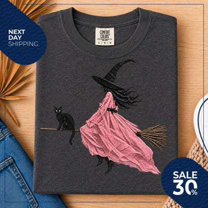 May include: A charcoal gray crew neck t-shirt featuring a graphic print of a figure in a black witch hat and a flowing pink dress riding a broomstick, accompanied by a black cat. The inner neck label reads Comfort Colors size L G G.