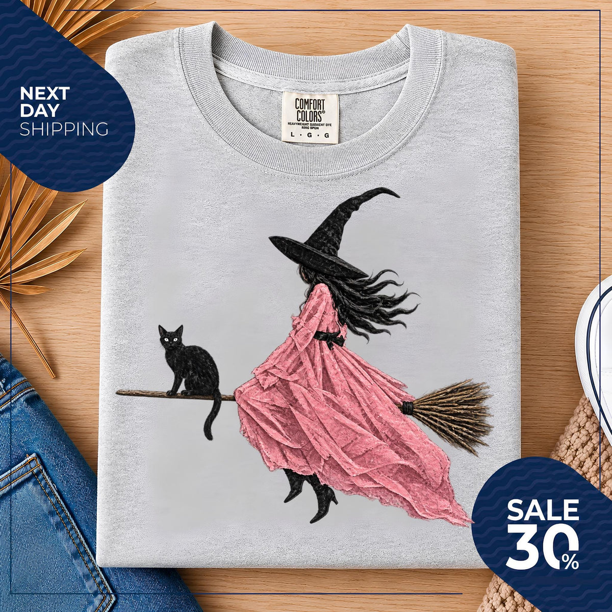 May include: A light gray crew neck t-shirt featuring a graphic illustration of a witch in a long pink dress and a black pointed hat riding a broomstick with a black cat sitting on the handle. The shirt has a Comfort Colors brand label at the neck.