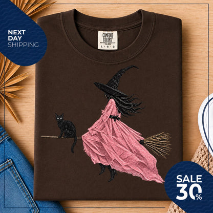 May include: A dark brown t-shirt featuring a graphic illustration of a figure in a black witch hat and a flowing pink dress riding a broomstick, accompanied by a black cat. The shirt has a Comfort Colors brand label at the inner neckline.