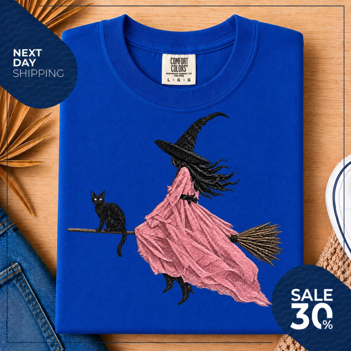May include: A royal blue t-shirt featuring a graphic illustration of a witch wearing a black hat and a flowing pink dress, riding a broomstick with a black cat perched on the handle. The shirt has a Comfort Colors brand label at the neck.