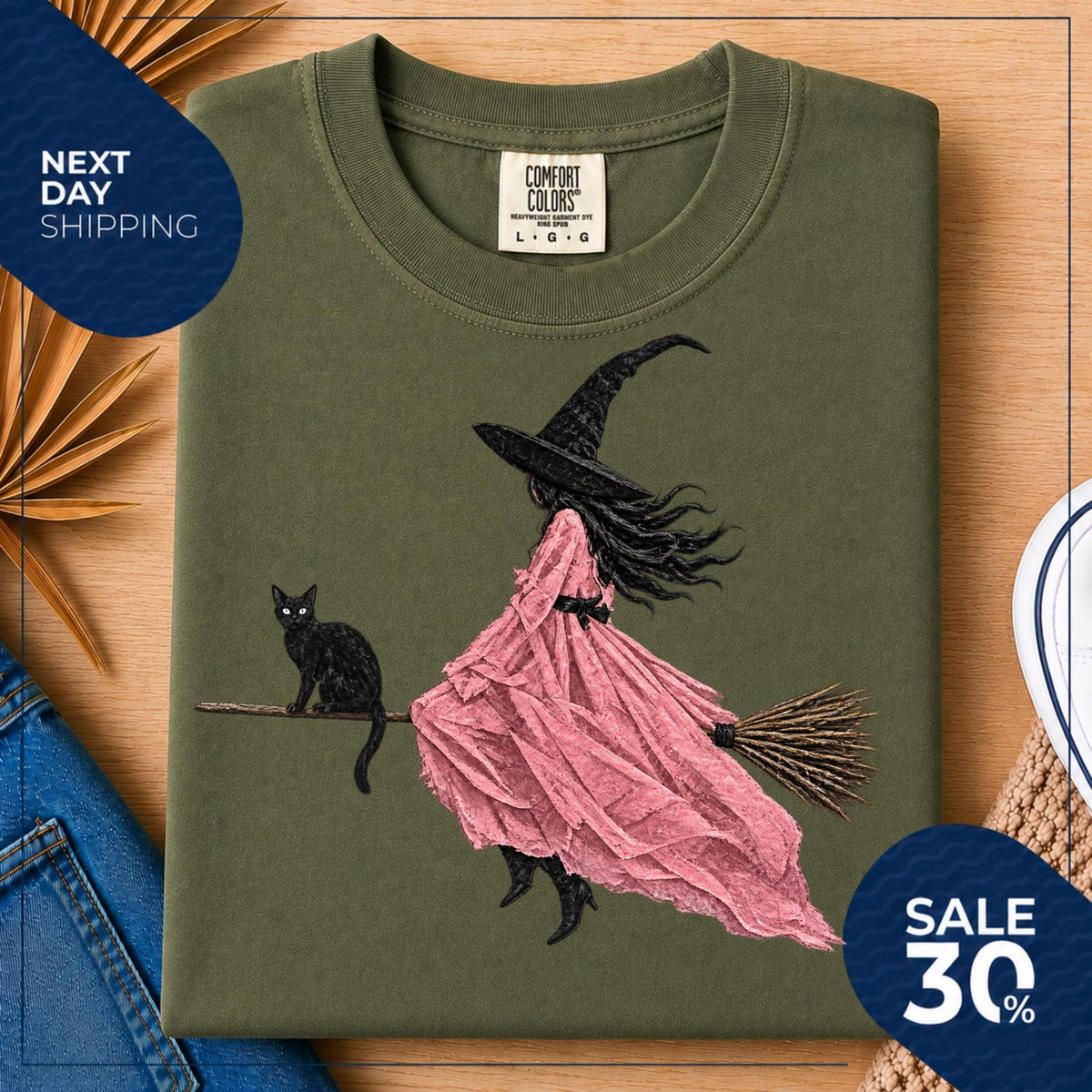 May include: An olive green crew neck t-shirt featuring a graphic illustration of a figure wearing a black witch hat and a flowing pink dress, riding a broomstick with a black cat sitting on the handle. The shirt has a Comfort Colors brand label.