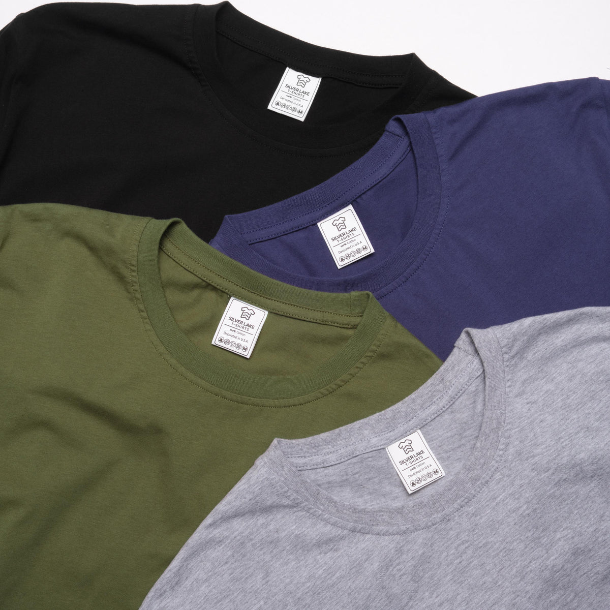 May include: A collection of four crew neck t-shirts in black, navy blue, olive green, and heather gray. Each shirt features a white interior neck label with the text Silver Lake T-Shirts, size M, and various laundry care icons printed on the fabric.