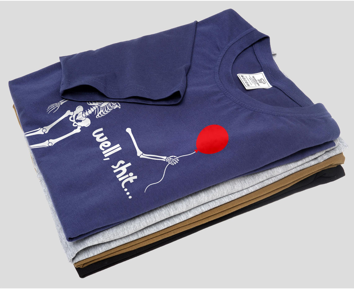 May include: A navy blue t-shirt featuring a white graphic of a skeleton holding a red balloon with the text "well, shit..." printed in white. The t-shirt is folded on top of a stack of other t-shirts in gray, tan, and black colors.