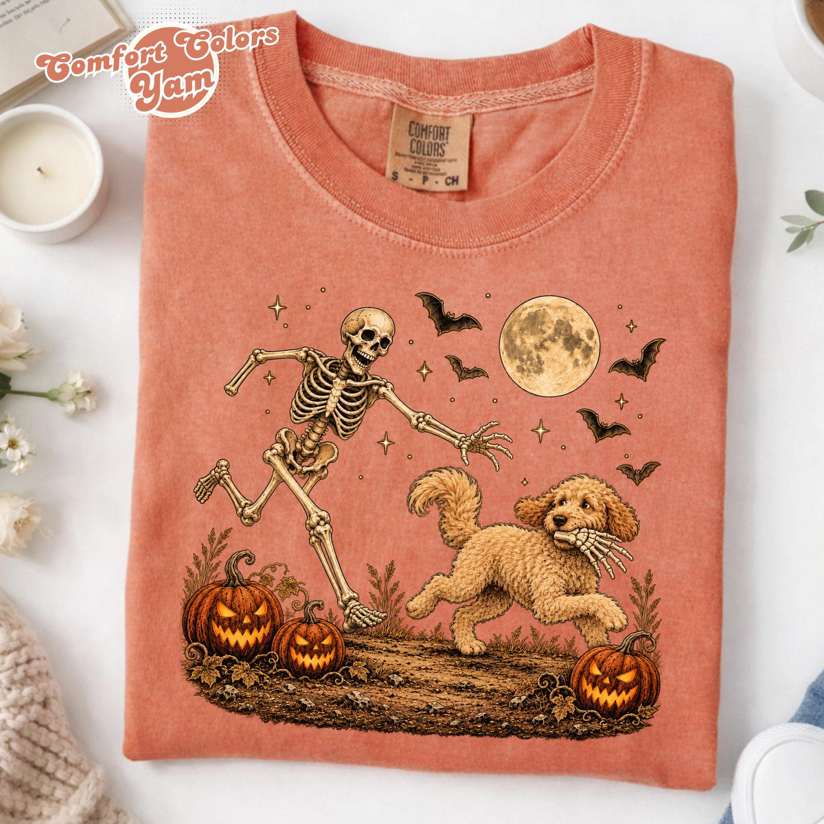 May include: A coral-colored t-shirt featuring a graphic print of a skeleton chasing a golden-colored dog. The design includes three jack-o'-lanterns, a full moon, and several bats. The shirt has a Comfort Colors brand label at the inner neckline.