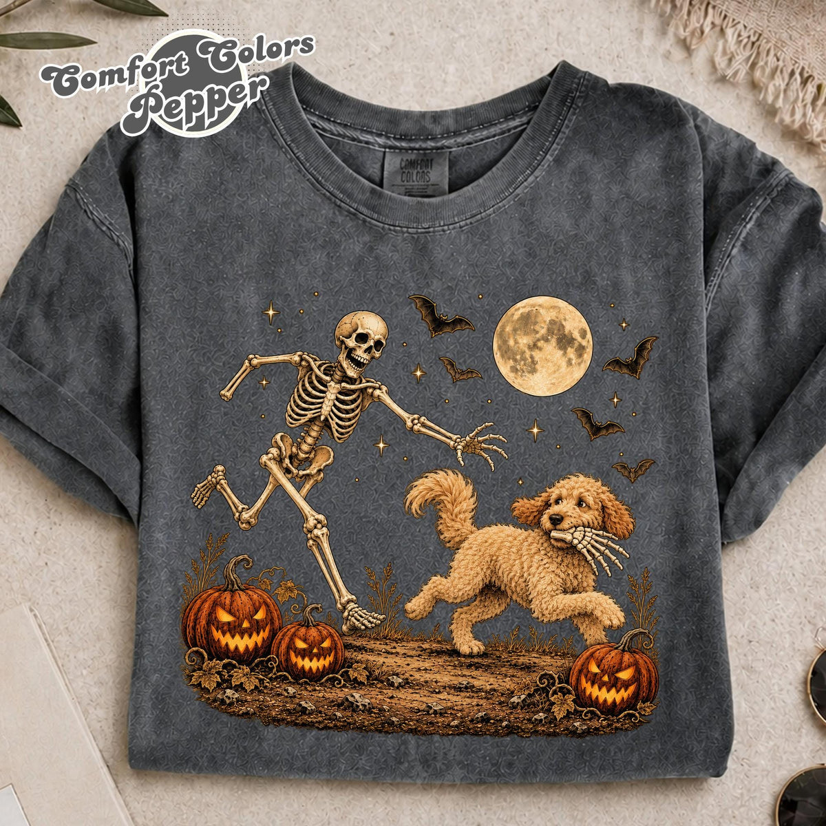 May include: A charcoal gray t-shirt featuring a graphic of a skeleton chasing a golden-colored dog that is holding a skeletal hand in its mouth. The scene includes two jack-o'-lanterns, a full moon, and flying bats against a dark background.