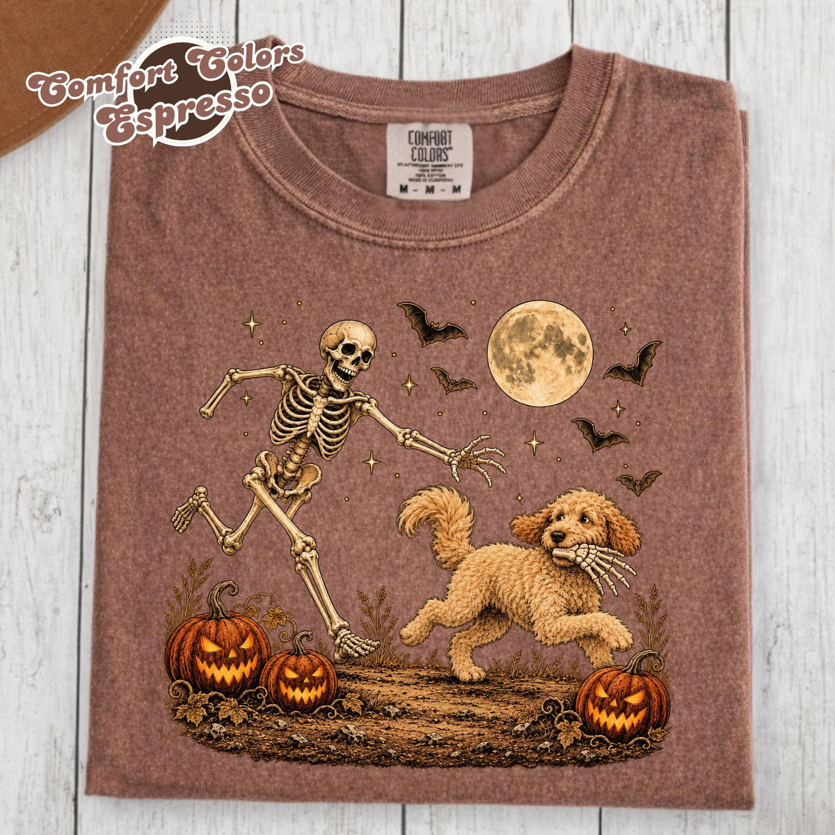 May include: A brown t-shirt featuring a graphic of a skeleton running after a golden doodle dog holding a skeletal hand in its mouth. The scene includes two jack-o'-lanterns, a full moon, and flying bats against a dark, textured background.
