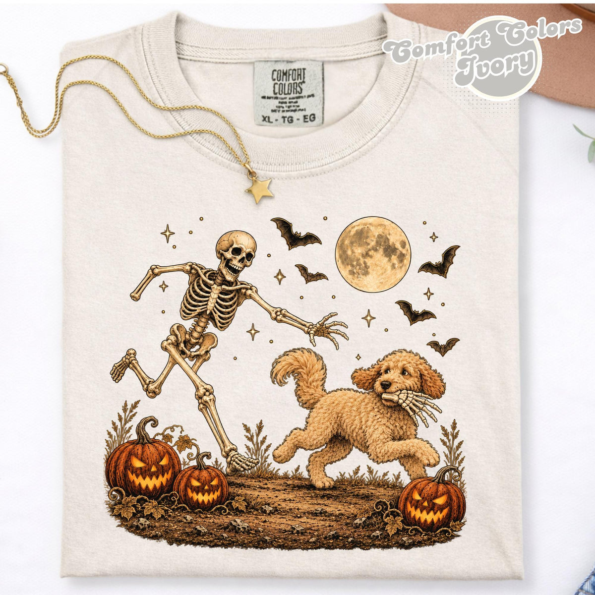May include: An ivory t-shirt featuring a graphic of a running skeleton chasing a golden doodle dog that is holding a skeletal hand in its mouth. The scene includes three jack-o'-lanterns, bats, and a full moon against a starry night sky.