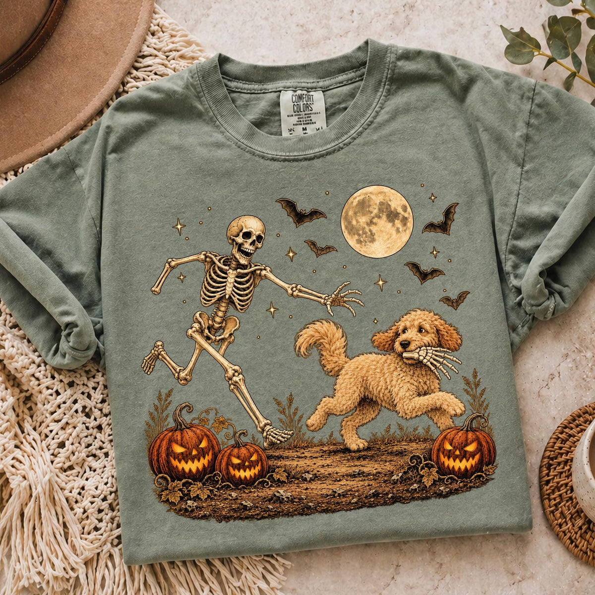 May include: A sage green t-shirt featuring a graphic of a running skeleton chasing a golden doodle dog that is holding a skeletal hand in its mouth. The scene includes two jack-o'-lanterns, a full moon, and flying bats against a dark sky.