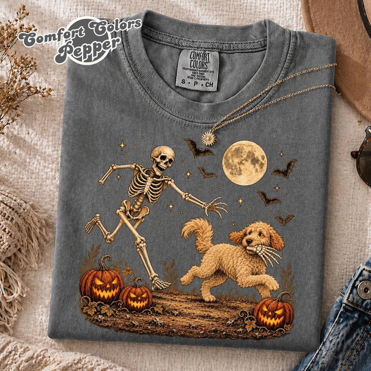 May include: A charcoal gray t-shirt featuring a graphic of a skeleton chasing a golden-colored dog that is holding a skeletal hand in its mouth. The design includes a full moon, bats, stars, and three jack-o'-lanterns on a dirt ground.