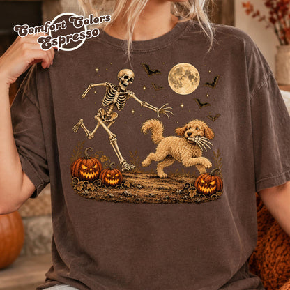 May include: A dark brown t-shirt featuring a graphic of a skeleton chasing a golden-colored dog that is holding a skeletal hand in its mouth. The scene includes two jack-o'-lanterns, a full moon, and flying bats. Text reads Comfort Colors Espresso.