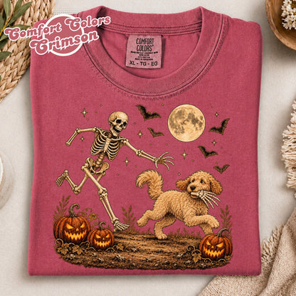 May include: A crimson t-shirt featuring a graphic of a skeleton running alongside a golden-colored dog that is holding a skeletal hand in its mouth. The scene includes two jack-o'-lanterns, a full moon, and several bats against a dark ground.