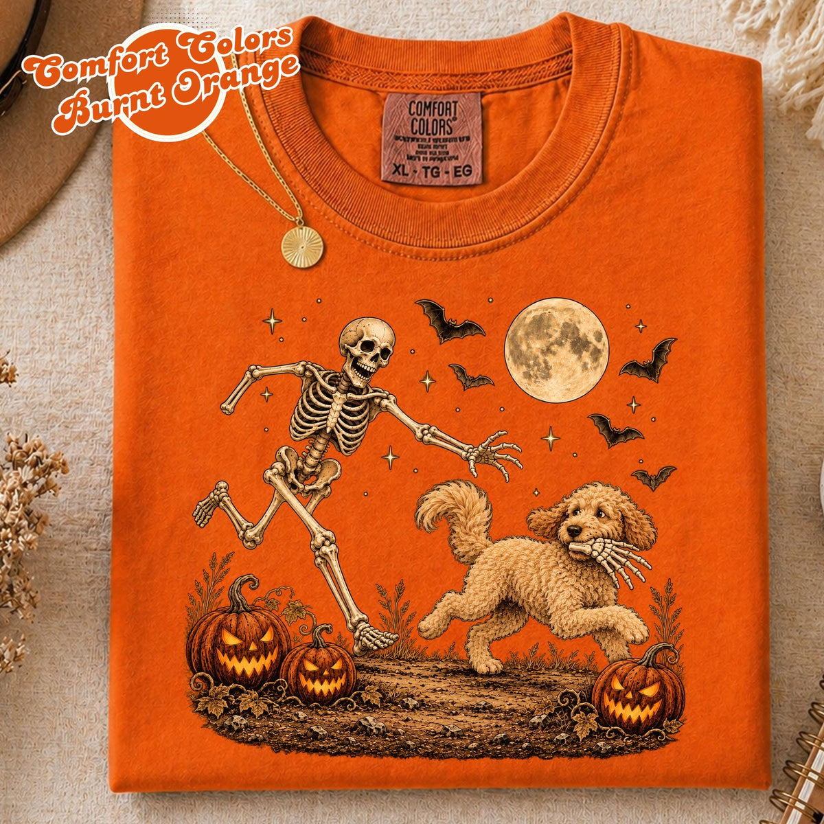 May include: A burnt orange t-shirt featuring a graphic of a running skeleton and a golden doodle dog carrying a skeletal hand in its mouth. The design includes two jack-o'-lanterns, a full moon, and flying bats against a dark ground.