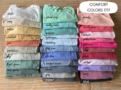 May include: A collection of folded Comfort Colors 1717 t-shirts arranged in three vertical stacks. The shirts display a wide variety of colors, each labeled with text including white, ivory, butter, mustard, yam, sandstone, bay, light green, blue spruce, moss, island reef, chalky mint, seafoam, hydrangea, chambray, blue jean, flo blue, washed denim, denim, gray, pepper, peachy, crunch berry, blossom, watermelon, heliconia, brick, chili, orchid, violet, berry, and black.