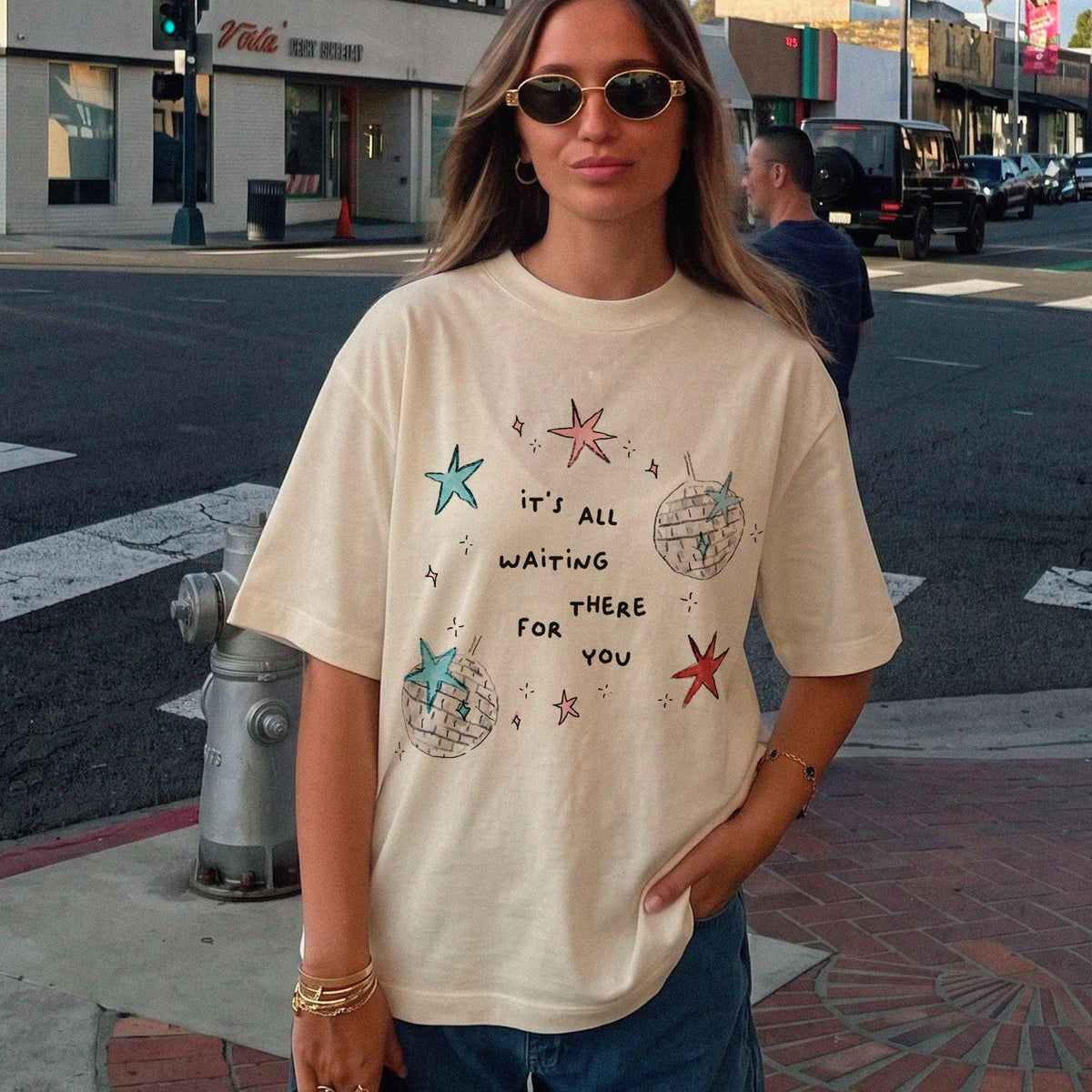 May include: A cream-colored short-sleeve t-shirt featuring a graphic print of two disco balls, several stars in red and blue, and the text "it's all waiting there for you" printed in black lettering across the center of the chest.