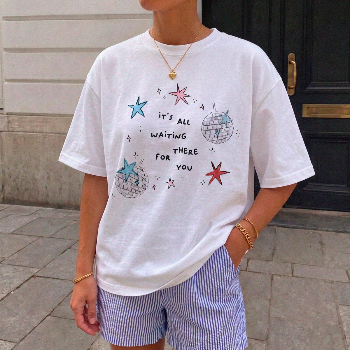 May include: A white short-sleeved t-shirt featuring a graphic print of two disco balls and several stars in blue, pink, and red. The text "it's all waiting for you" is printed in black in the center. The shirt is paired with blue and white striped shorts.