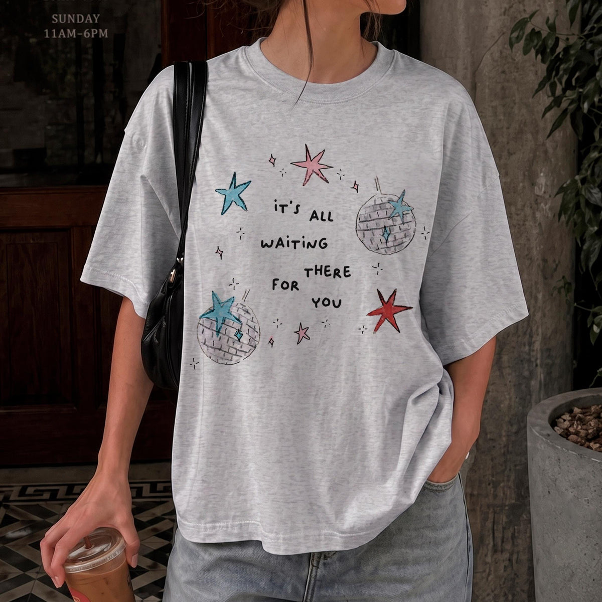 May include: A light gray short-sleeve t-shirt featuring a graphic print with the text 'IT'S ALL WAITING THERE FOR YOU' surrounded by illustrations of two disco balls and several stars in shades of blue, pink, and red on the front.