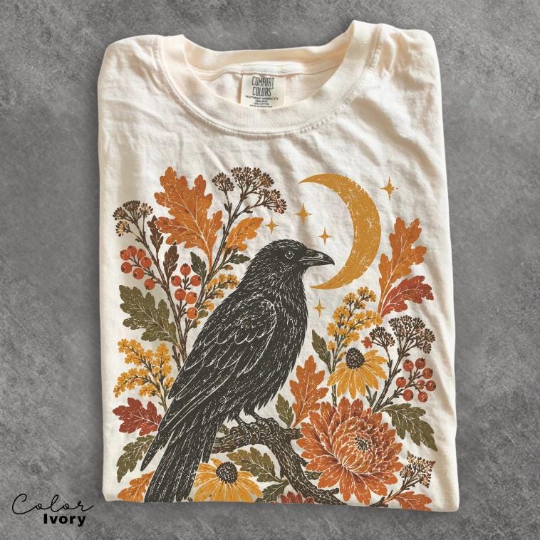 May include: An ivory t-shirt featuring a graphic print of a black crow perched on a branch. The design includes orange and yellow autumn leaves, berries, flowers, and a crescent moon with small stars. The neck label reads Comfort Colors.
