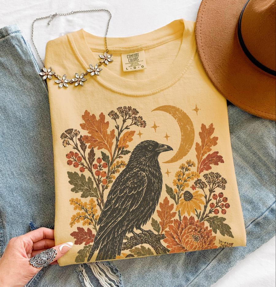 May include: A yellow t-shirt featuring a graphic print of a black crow perched on a branch, surrounded by autumn leaves, berries, flowers, and a crescent moon. The shirt has a Comfort Colors label in size large and is displayed with a brown hat.