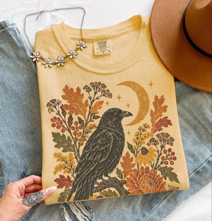 May include: A yellow t-shirt featuring a graphic print of a black crow perched on a branch, surrounded by autumn leaves, berries, flowers, and a crescent moon. The shirt has a Comfort Colors label in size large and is displayed with a brown hat.