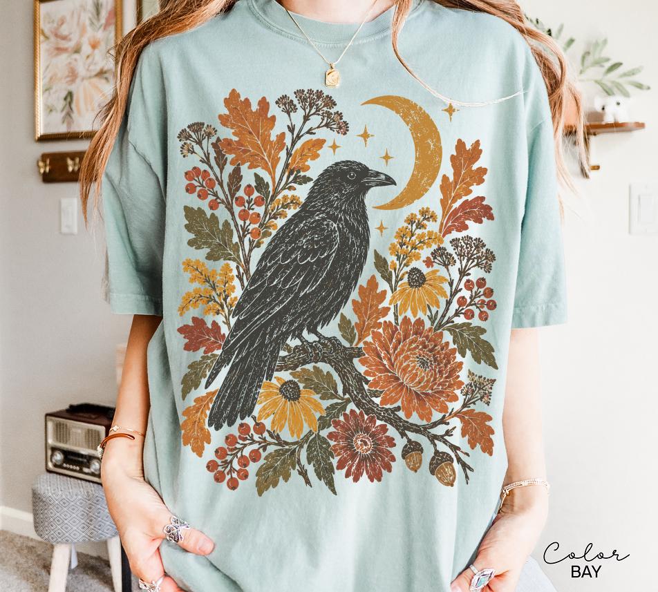 May include: A light blue short-sleeve t-shirt featuring a graphic print of a black crow perched on a branch surrounded by orange and yellow autumn leaves, berries, flowers, and a crescent moon. The text Color Bay is printed in the bottom right corner.