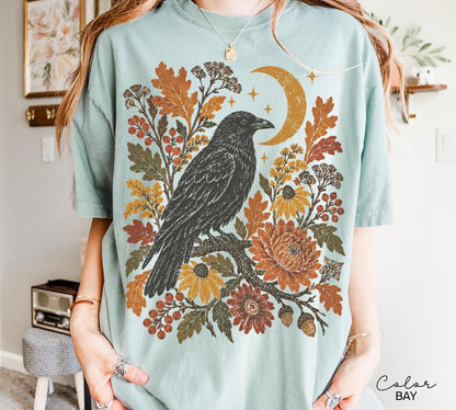 May include: A light blue short-sleeve t-shirt featuring a graphic print of a black crow perched on a branch surrounded by orange and yellow autumn leaves, berries, flowers, and a crescent moon. The text Color Bay is printed in the bottom right corner.