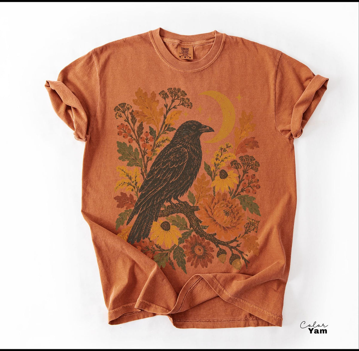 May include: A burnt orange short-sleeve t-shirt featuring a graphic print of a black crow perched on a branch surrounded by autumn-toned flowers, leaves, and a crescent moon. The shirt has a crew neckline and a relaxed, casual fit.