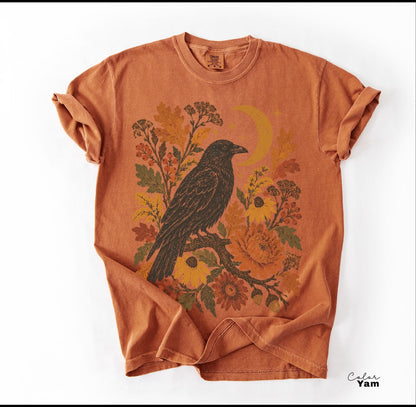 May include: A burnt orange short-sleeve t-shirt featuring a graphic print of a black crow perched on a branch surrounded by autumn-toned flowers, leaves, and a crescent moon. The shirt has a crew neckline and a relaxed, casual fit.