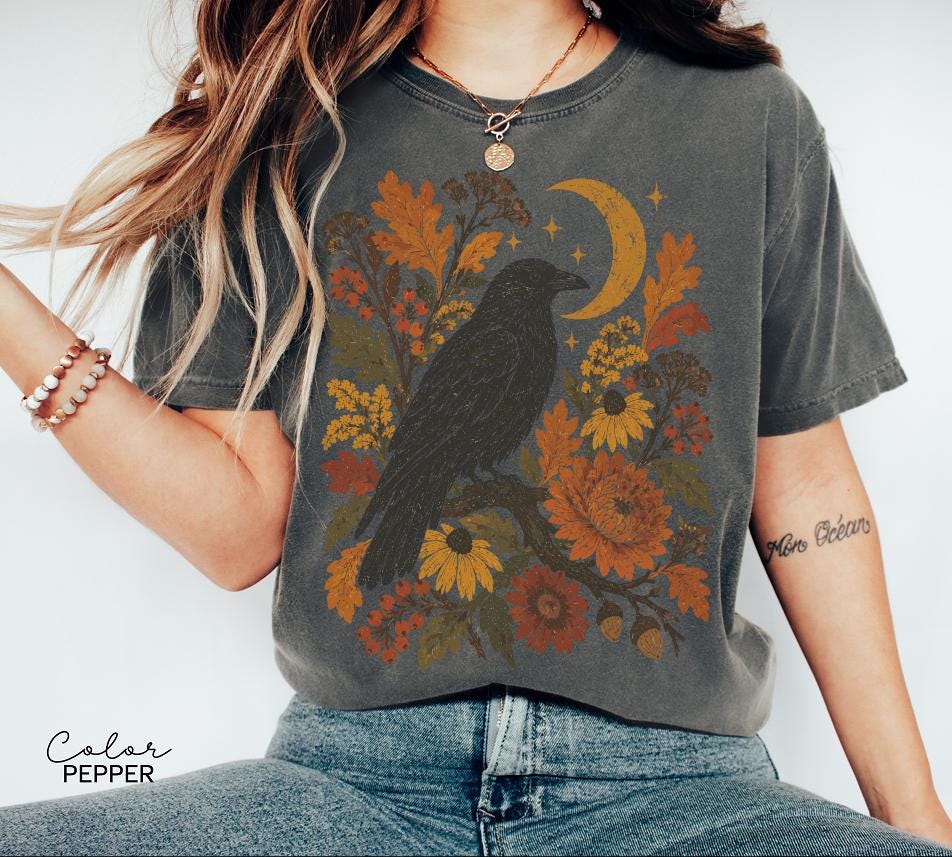May include: A charcoal gray t-shirt featuring a graphic print of a black raven perched on a branch surrounded by autumn-toned flowers, leaves, and a crescent moon. The text "Color Pepper" is printed in the bottom left corner of the image.