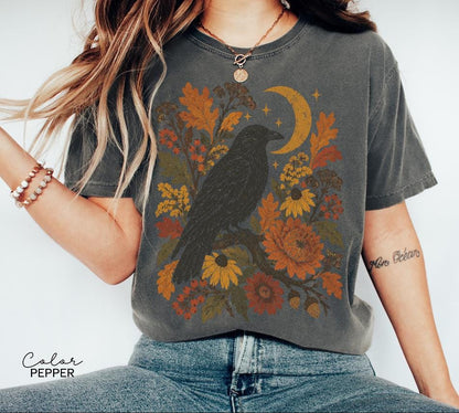 May include: A charcoal gray t-shirt featuring a graphic print of a black raven perched on a branch surrounded by autumn-toned flowers, leaves, and a crescent moon. The text "Color Pepper" is printed in the bottom left corner of the image.