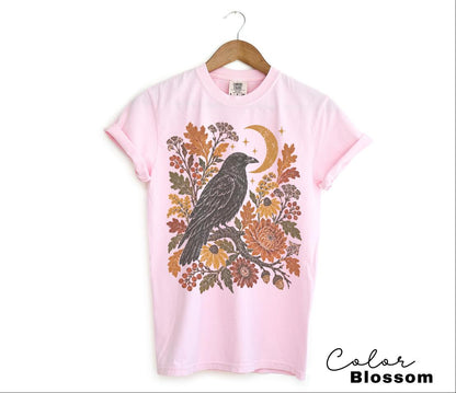 May include: A light pink t-shirt featuring a graphic print of a black crow perched on a branch, surrounded by orange and brown autumn leaves, flowers, berries, and a crescent moon. The shirt is displayed on a wooden hanger against a white background.