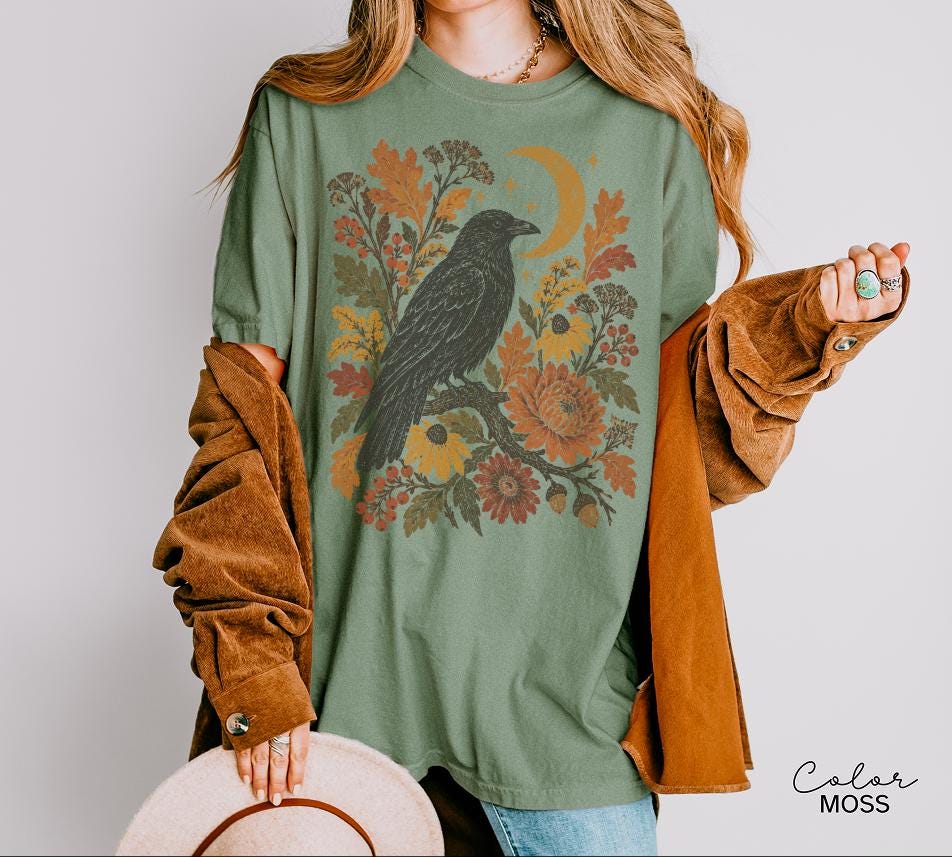 May include: A moss green t-shirt featuring a graphic print of a black raven perched on a branch, surrounded by autumn-toned flowers, leaves, and a crescent moon. The shirt is layered under a brown corduroy button-down shirt and a wide-brimmed hat.